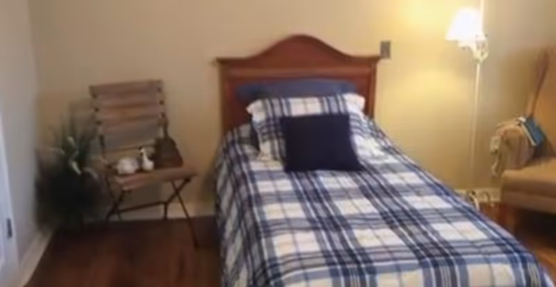 Small bedroom with a single bed covered in blue plaid bedding, wooden headboard, a chair and a floor lamp.