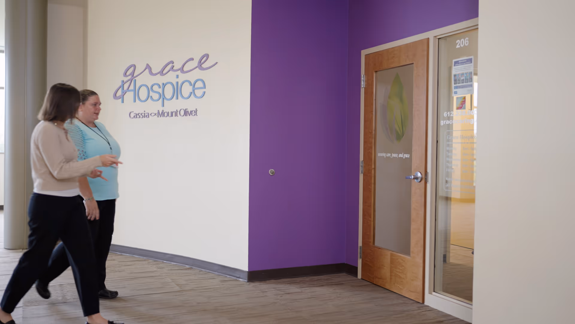 Two people walk through a Grace Hospice hallway with a purple wall and logo.