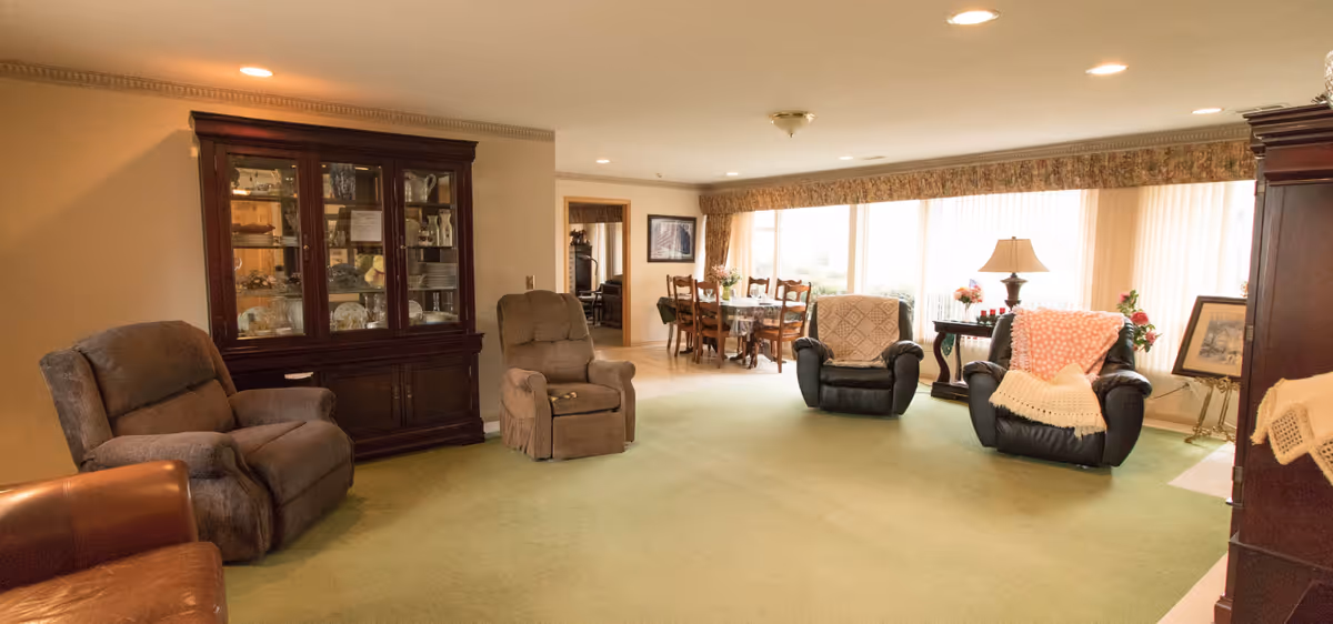 Cozy senior living lounge with recliners, a china cabinet, dining table, and large windows.