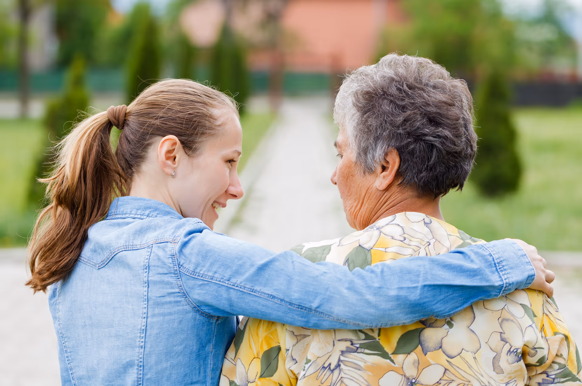 Two women walk arm-in-arm along a park path outdoors.