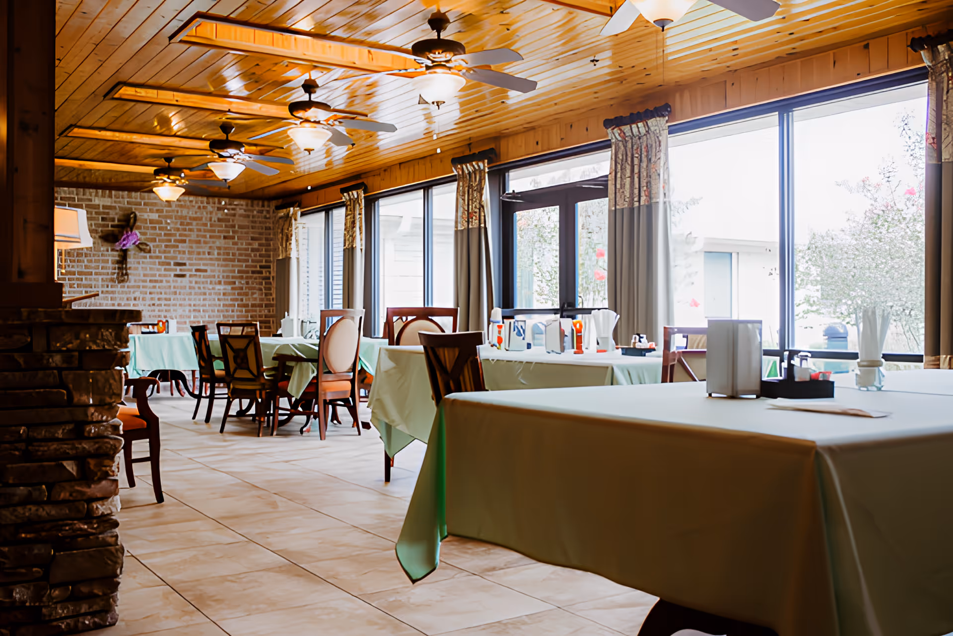 Sunlit dining room with tables covered in green tablecloths, wooden chairs, ceiling fans, and large windows.
