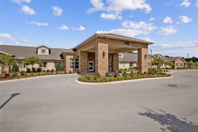 Front entrance of a single-story brick senior living facility with a covered porte-cochère, landscaped island, and circular driveway under a blue sky.