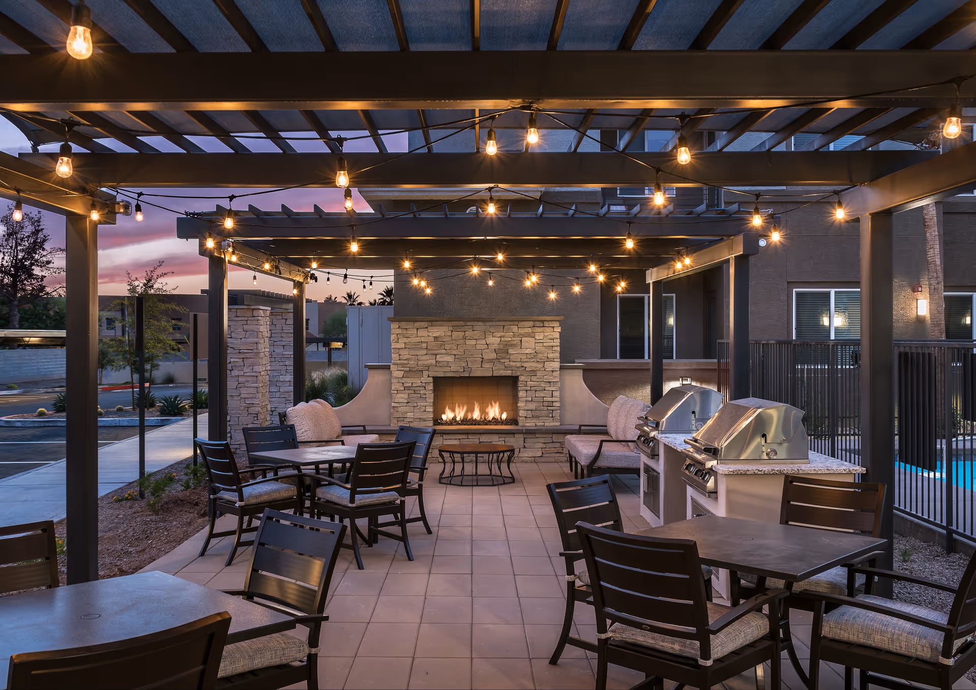 Covered outdoor patio with string lights, dining tables, grills, and a stone fireplace at dusk.