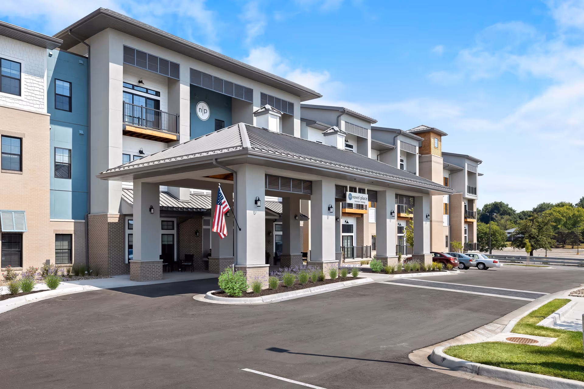 Modern senior living building with a covered entrance, flag, and cars in the parking lot.