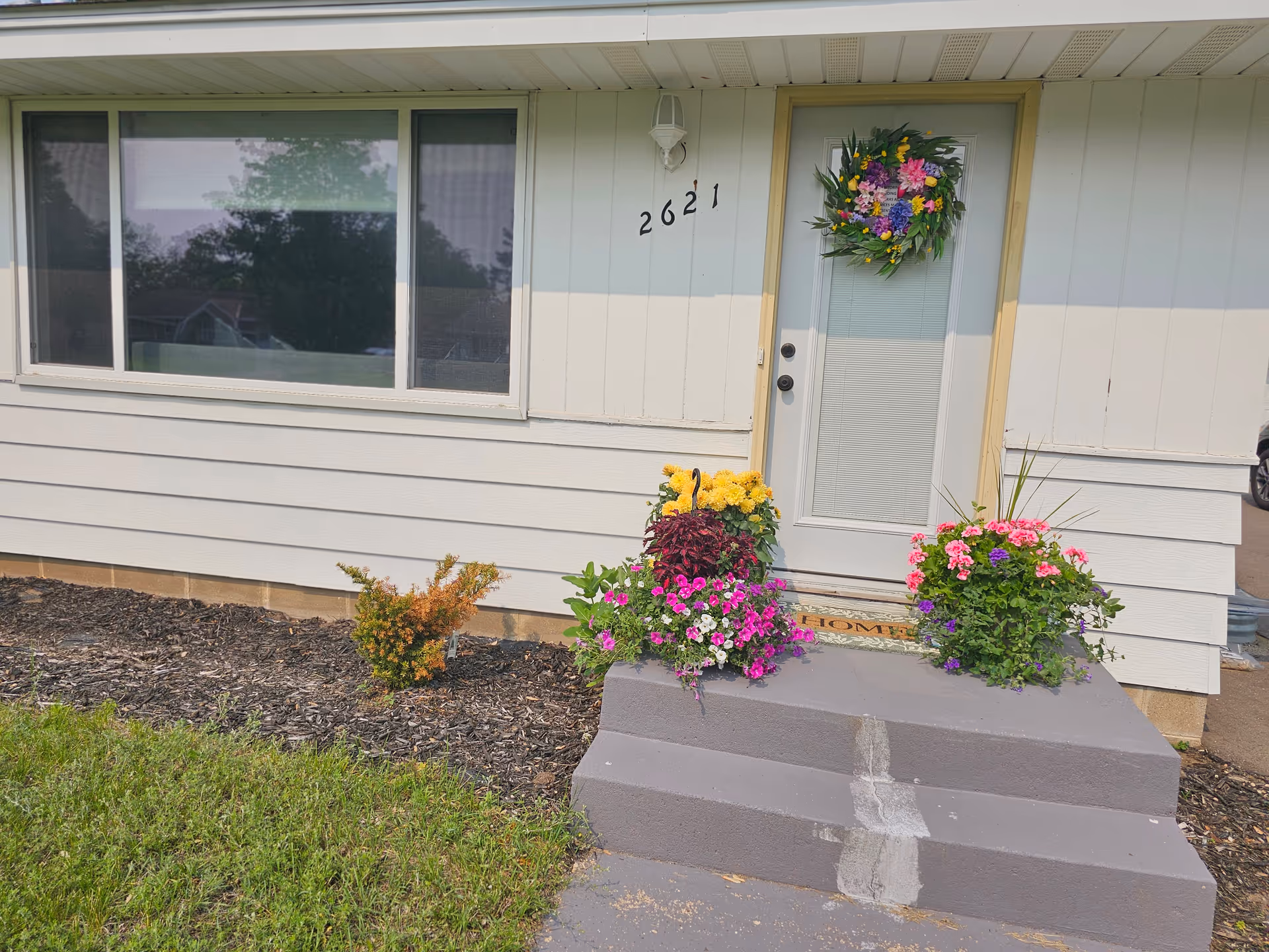 Front entrance of a light-siding building with steps, wreath, and colorful flower pots.