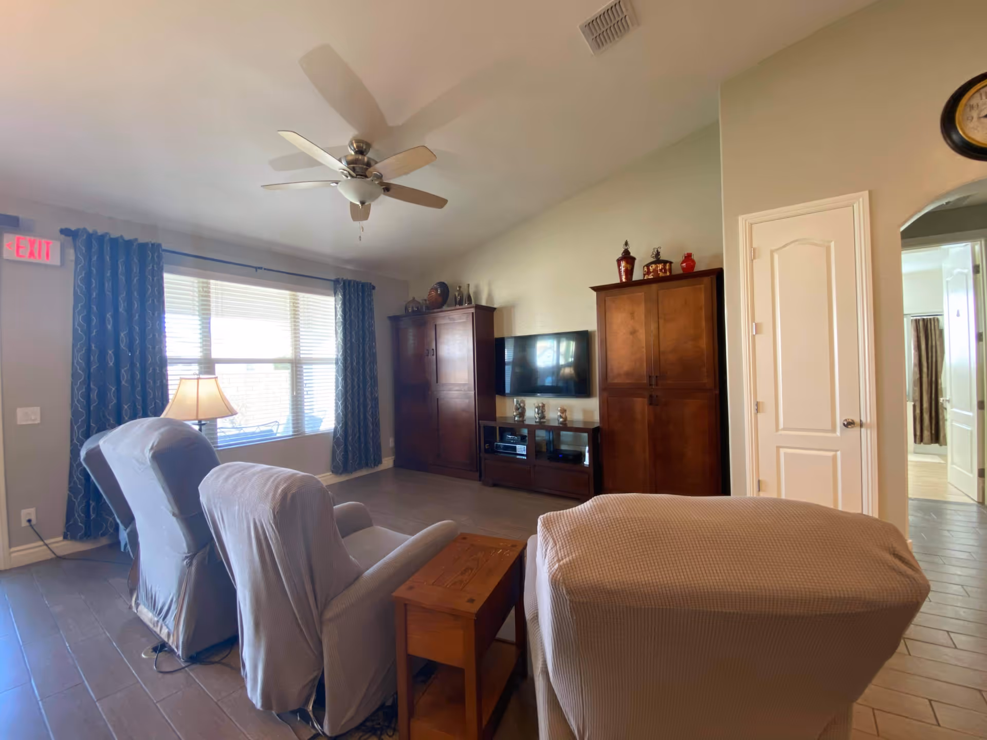 Spacious living room with recliners, a ceiling fan, windows with blue curtains, and an entertainment cabinet.