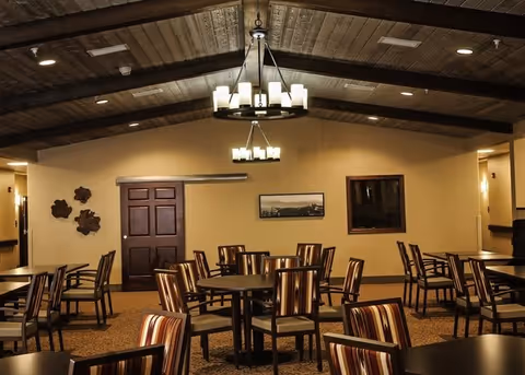 A warmly lit dining room with multiple round tables and striped cushioned chairs arranged neatly. The room features a high wooden ceiling with exposed beams and two modern chandeliers hanging down. The walls are beige with a dark wooden door, a framed picture, and decorative wall art. The carpet has a patterned design.