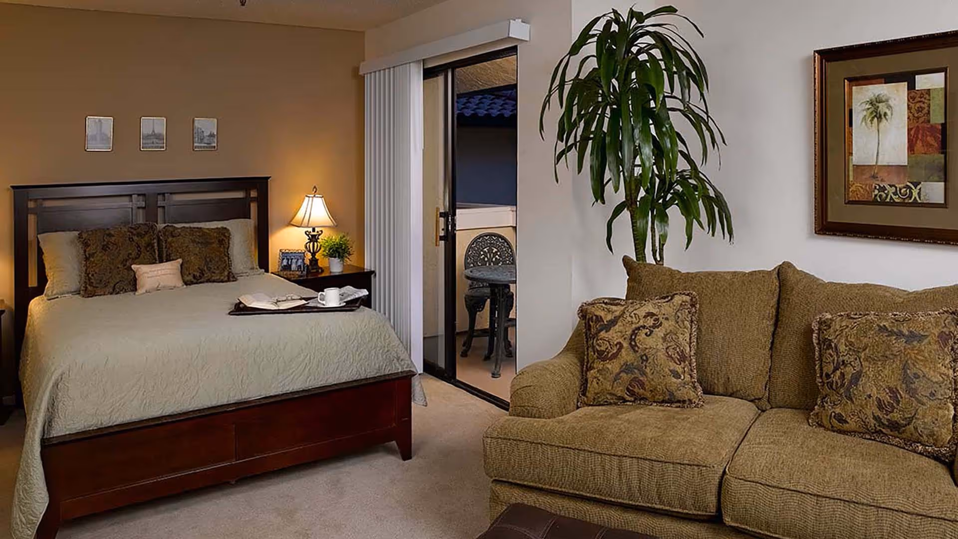 Furnished studio-style room with a bed, nightstand and lamp on the left and a couch and potted plant on the right with a sliding door to a small balcony.