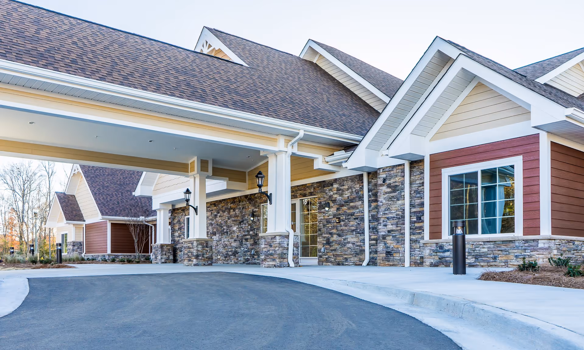 Exterior view of The Pearl at Five Forks senior living facility showing a covered entrance with stone and wood siding, large windows, and a paved driveway.