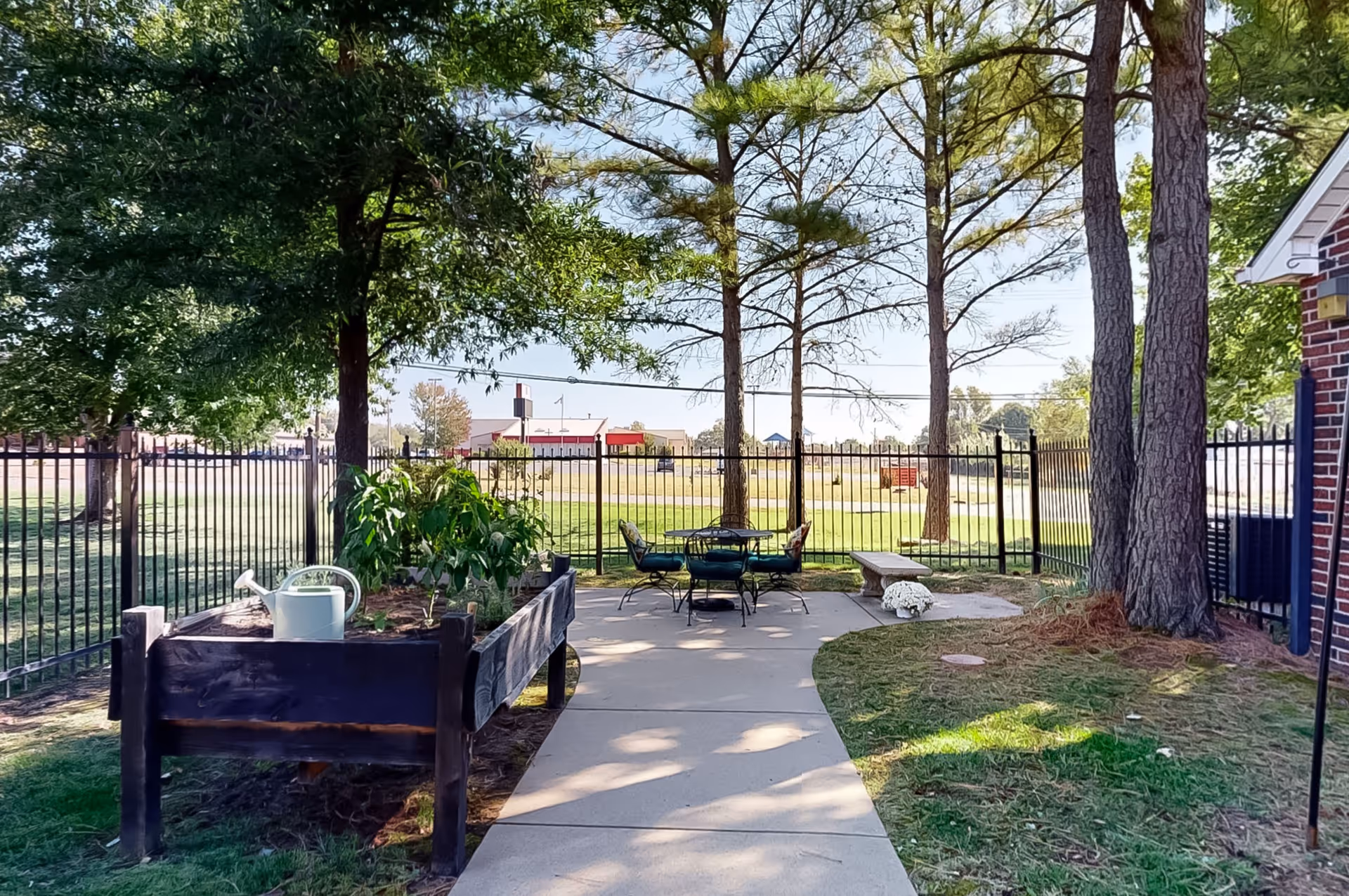 Outdoor courtyard with seating, a raised garden bed, and tall trees beside a fenced facility.