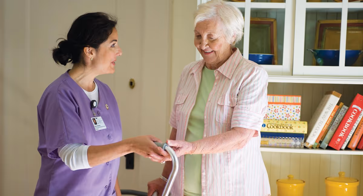 A female healthcare worker in a purple uniform assists an elderly woman using a walker inside a room with a white cabinet filled with cookbooks and yellow containers in the background.