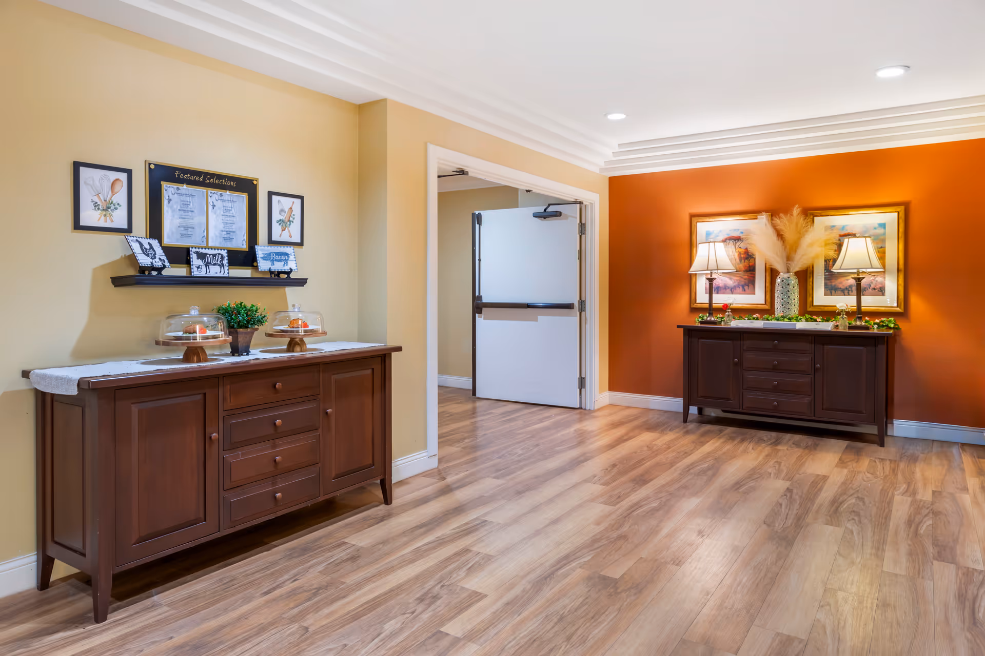 Interior hallway area with wooden flooring and two dark wooden sideboards against the walls. One sideboard is against a beige wall with framed pictures and a small plant with covered pastries on top. The other sideboard is against an orange wall with two framed pictures, two table lamps, and a vase with pampas grass. A white door with a push bar is open in the background.