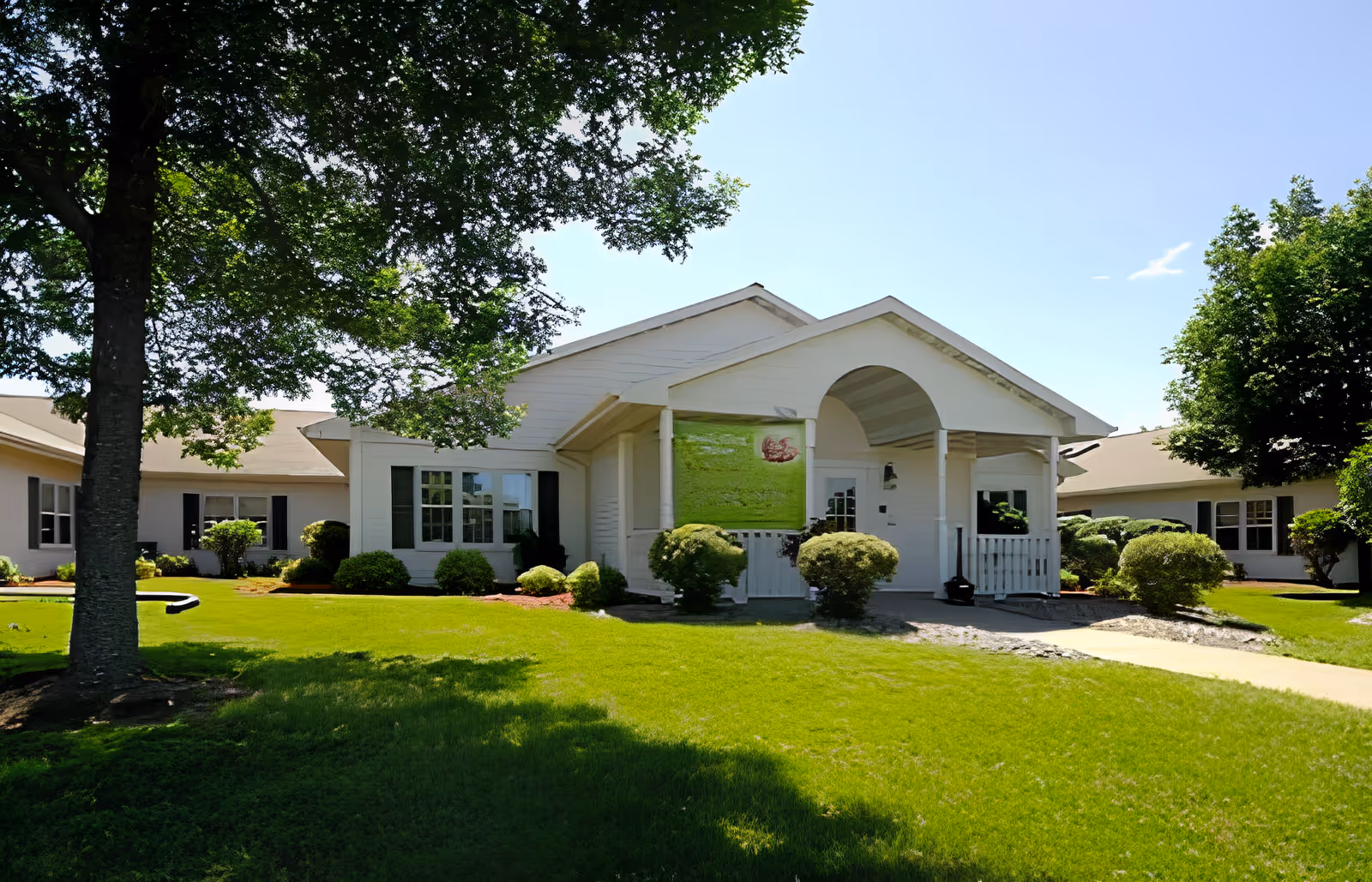 Front entrance of a single-story white senior living building with a covered porch, well-kept lawn and trees.