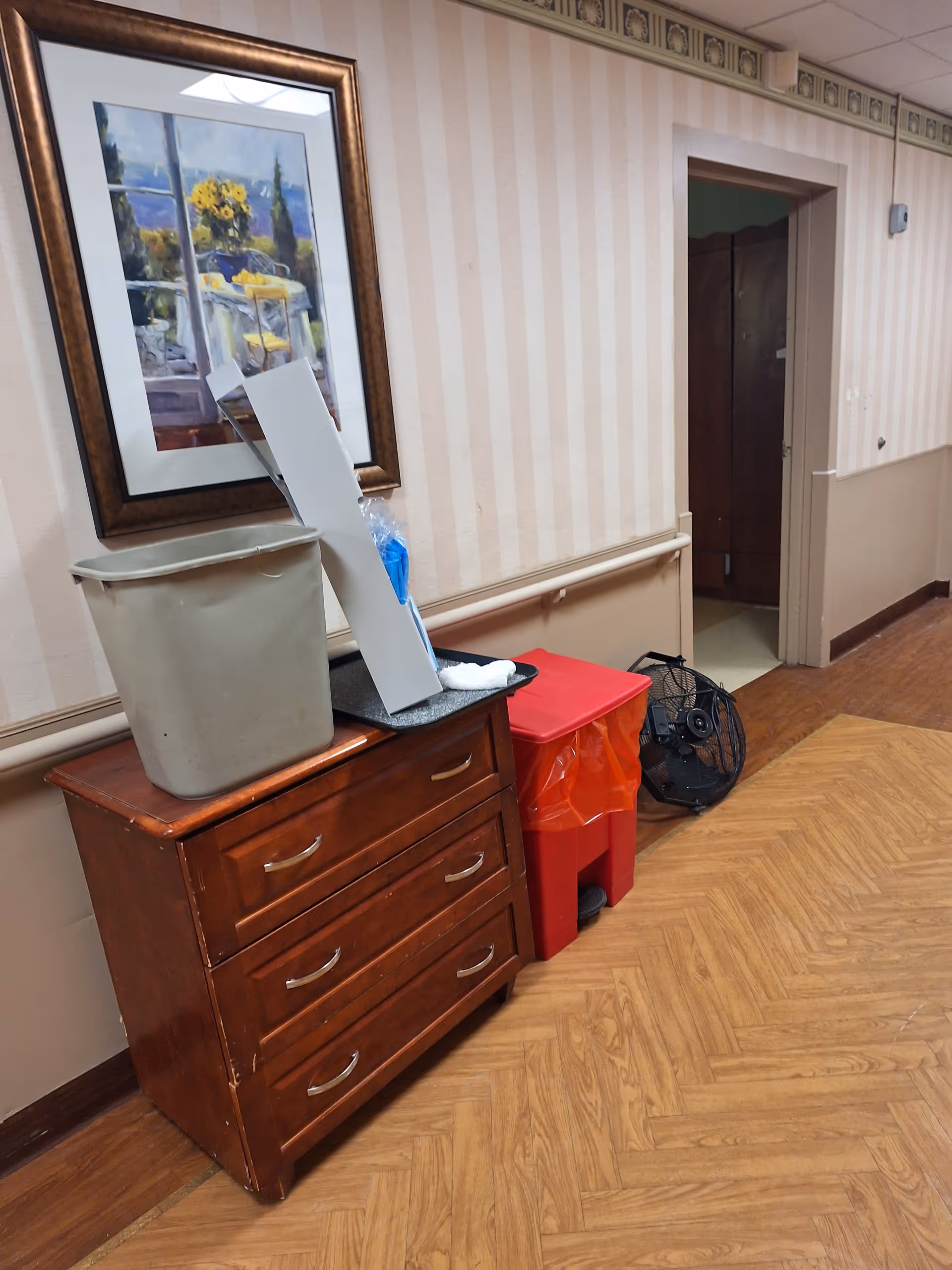 Indoor hallway of a senior living facility with a dresser, trash can, and a standing fan.