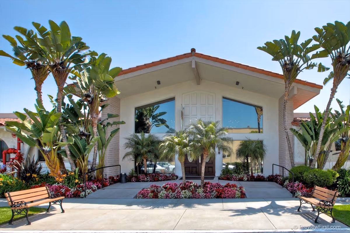 Front exterior view of Brittany House Residential Memory Care building with large windows, a central double door entrance, surrounded by palm trees, colorful flower beds, and two wooden benches on either side of the walkway.