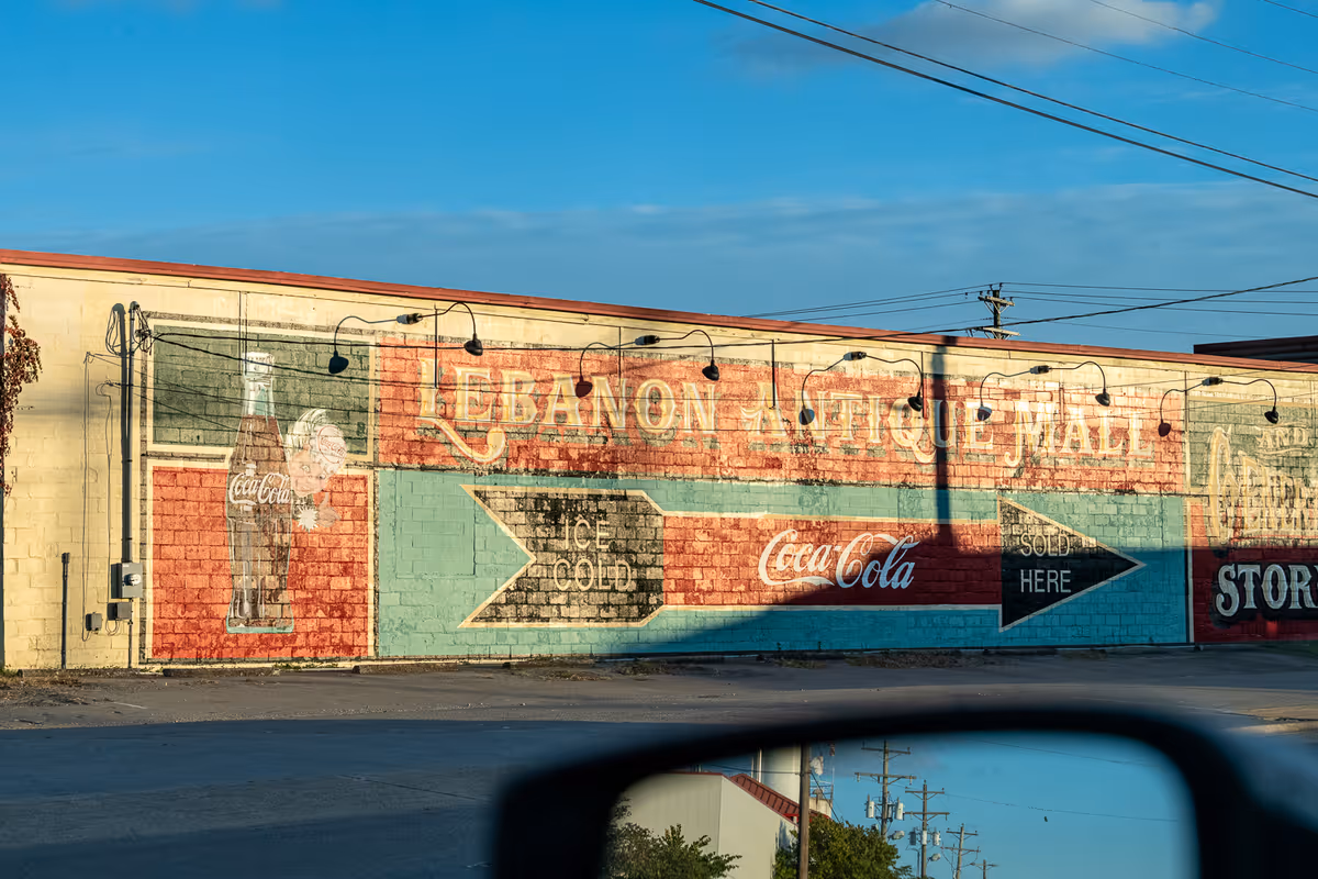 Sunlit painted brick exterior wall advertising 'Lebanon Antique Mall' and Coca-Cola with directional arrows, seen from a car with a side mirror in the foreground.