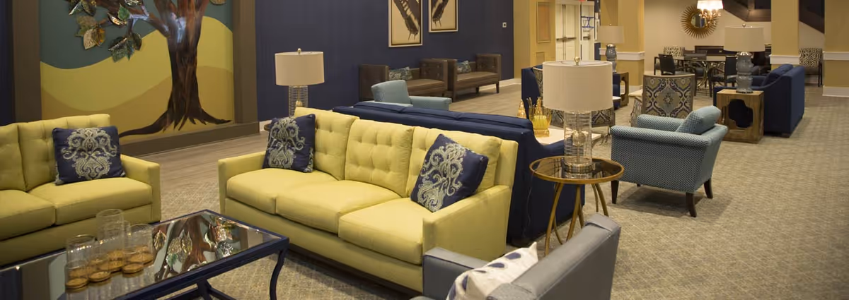 A spacious and well-lit living room area with multiple seating options including yellow and blue sofas, armchairs, and side tables with lamps. The room features a decorative wall with a tree mural and framed artwork, along with a carpeted floor and a glass coffee table with decorative items.