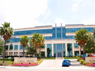 A modern glass-front building with palm trees and a blue car parked out front.