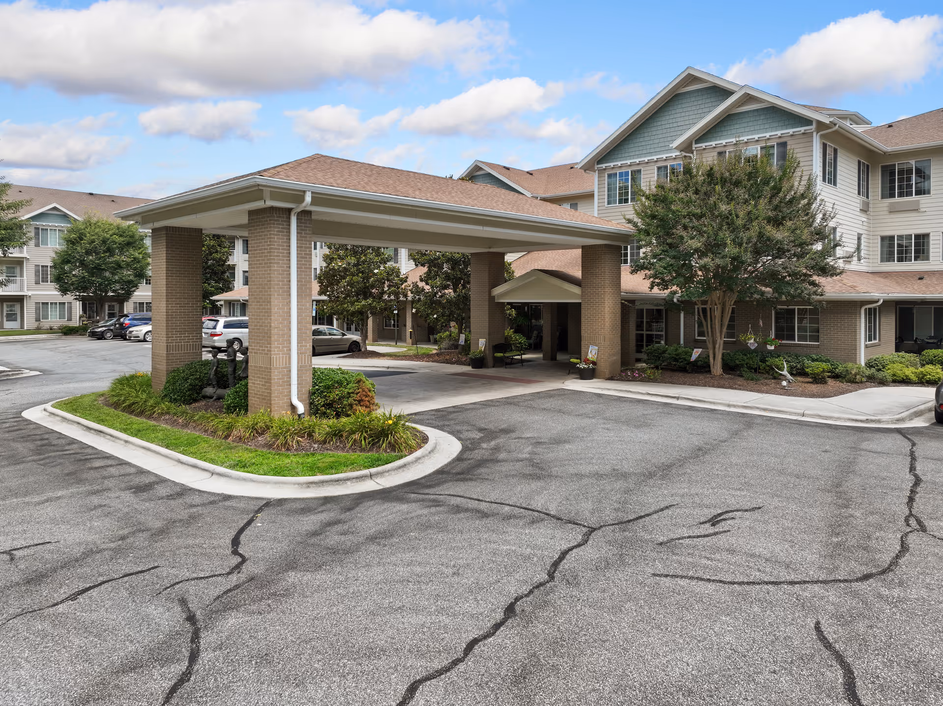 Covered porte-cochere entrance and driveway in front of a multi-story senior living building with landscaping and parked cars.