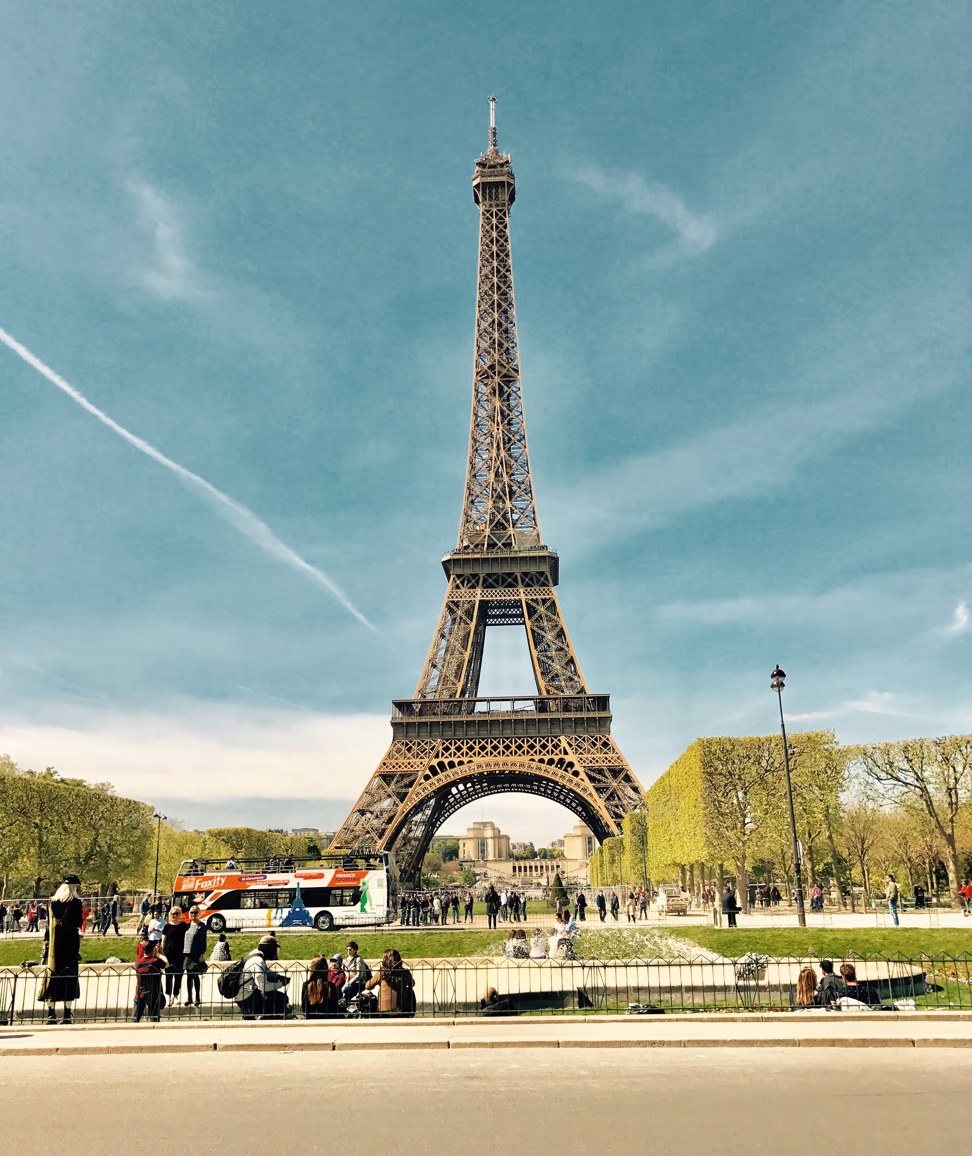 Crowds gather around the Eiffel Tower in a sunny Paris park.