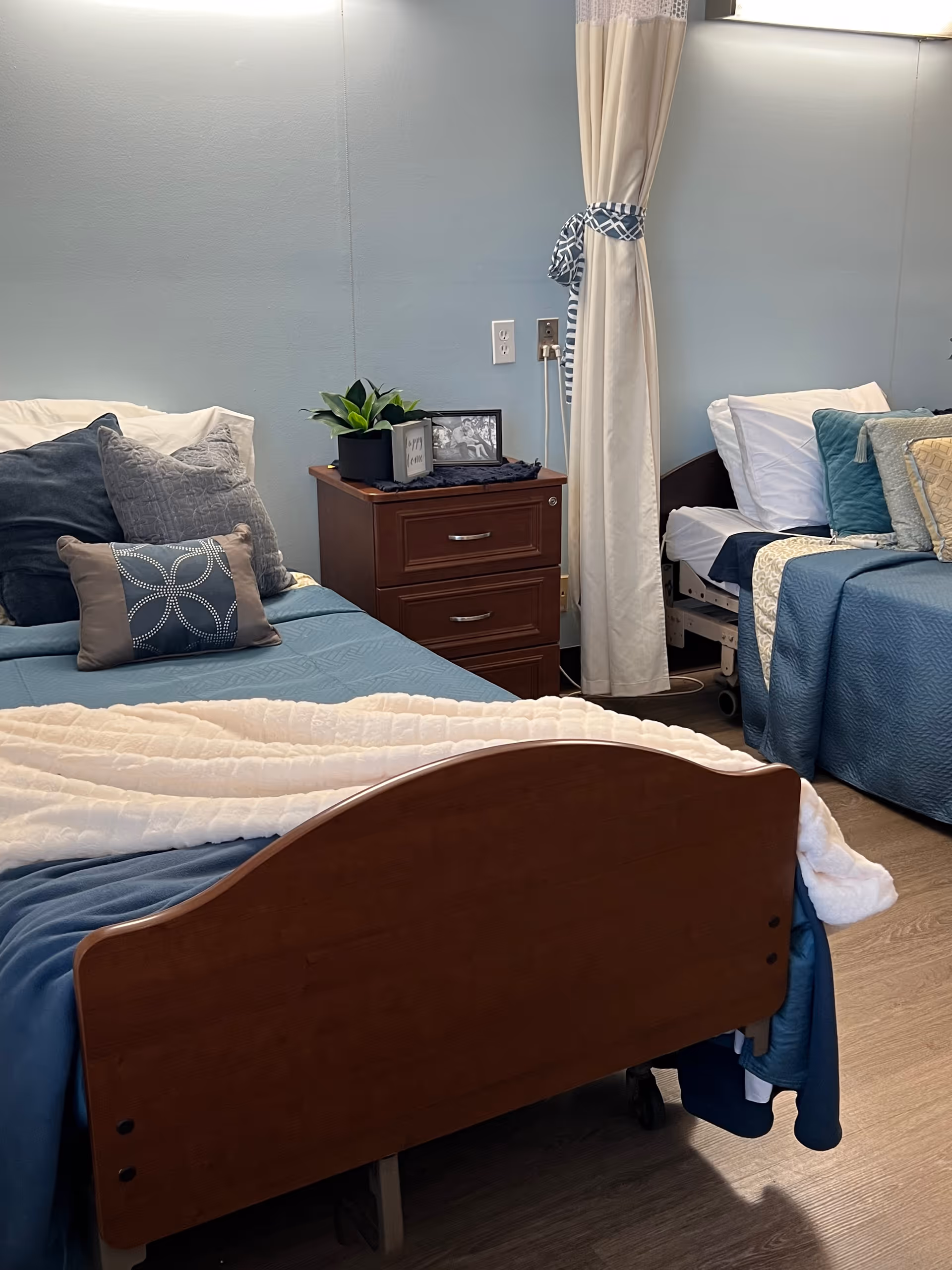 A room with two hospital-style beds, each made with blue bedding and multiple pillows. Between the beds is a wooden nightstand with two drawers, a small potted plant, and a framed photo. A privacy curtain is partially drawn between the beds. The walls are painted light blue and the floor has wood-like flooring.