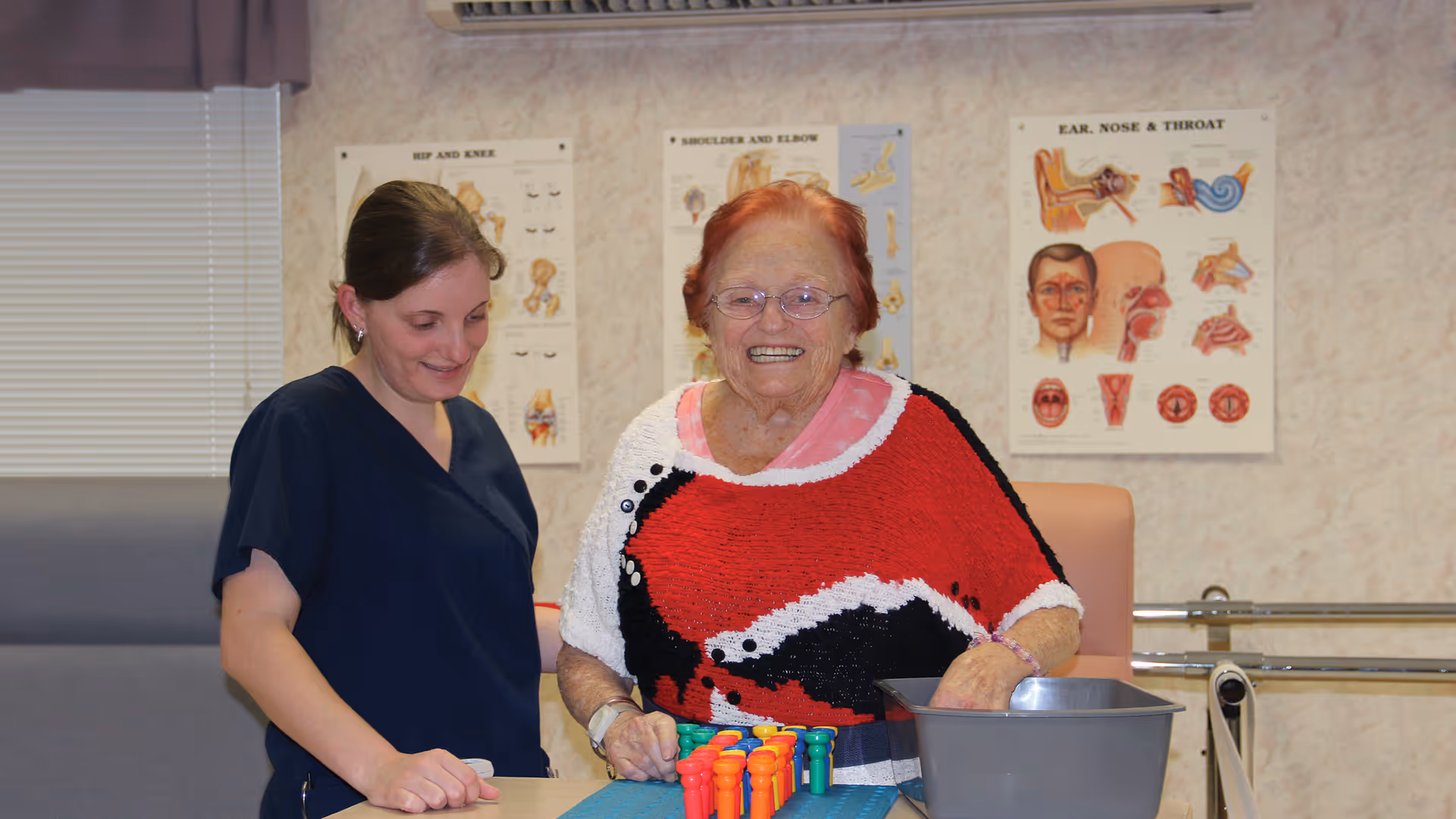 An elderly woman wearing glasses and a colorful sweater is smiling while standing next to a younger woman in navy scrubs. They are in a medical or therapy room with anatomical posters on the wall and a gray container with colorful pegs on the table in front of them.