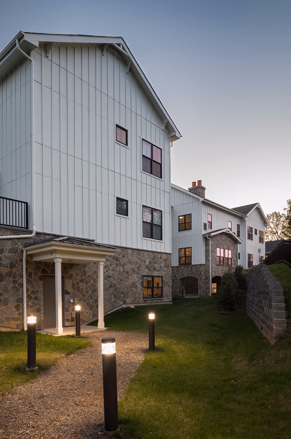 Exterior view of a multi-story residential building at dusk with white siding and stone accents. A pathway lined with illuminated bollard lights leads to a covered entrance. The sky is clear and the windows reflect the fading light.
