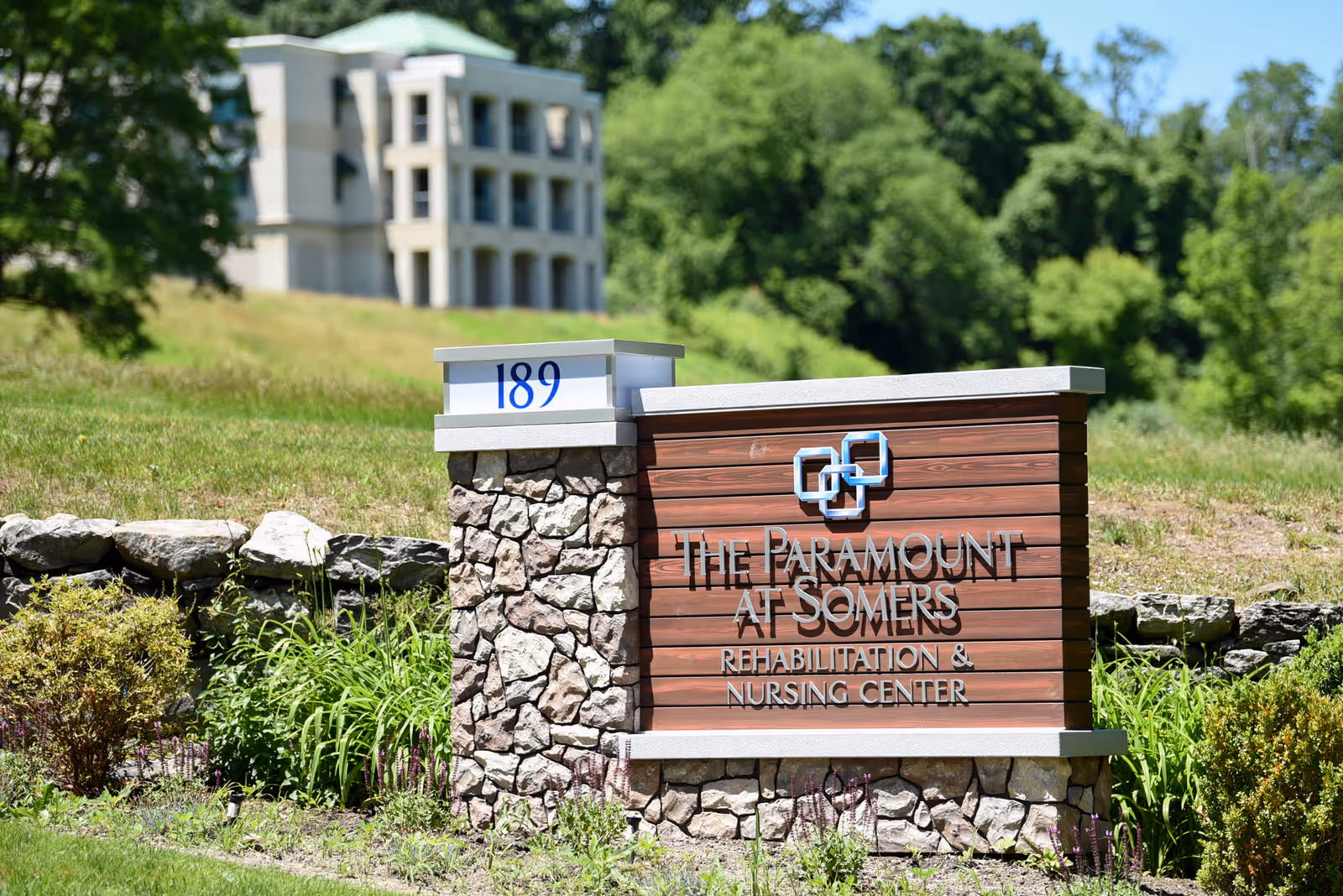 Outdoor view of a stone and wood sign for The Paramount at Somers Rehabilitation & Nursing Center with the number 189 on top. The sign is surrounded by greenery and landscaping, with a multi-story building and trees visible in the background under a clear blue sky.