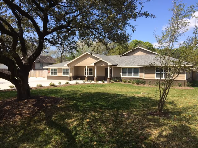 Single-story beige senior living facility with front entrance, manicured lawn, and surrounding trees.