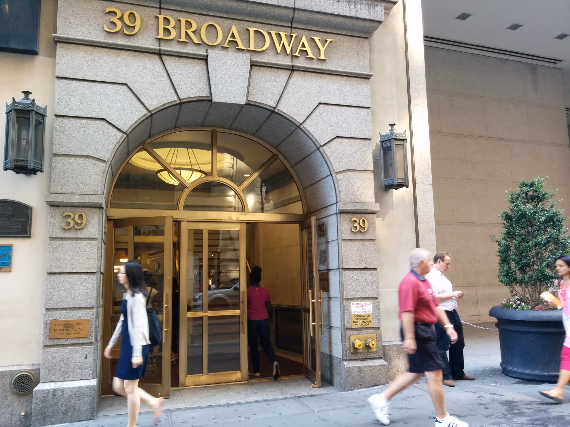 Golden glass doors entrance with arched stone facade, 39 Broadway, pedestrians nearby.