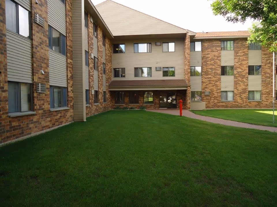 Exterior view of a multi-story brick and siding building with several windows, a green lawn in front, a paved walkway leading to the entrance, and a tree on the right side.