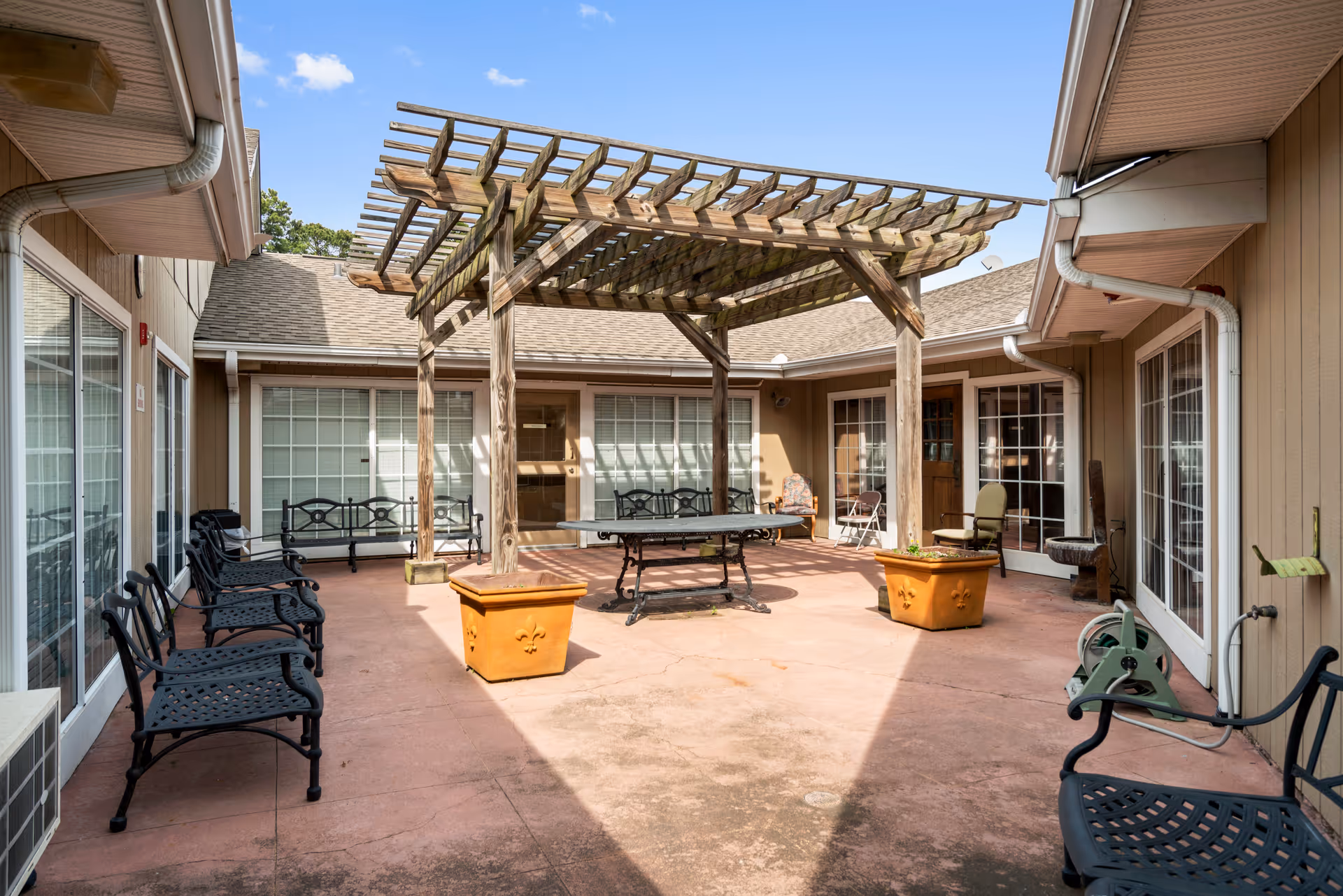 Outdoor courtyard area with a wooden pergola in the center, surrounded by several black metal benches and chairs. There are large windows and doors on the surrounding building walls, and two large orange planters near the pergola. The sky is clear with a few clouds.
