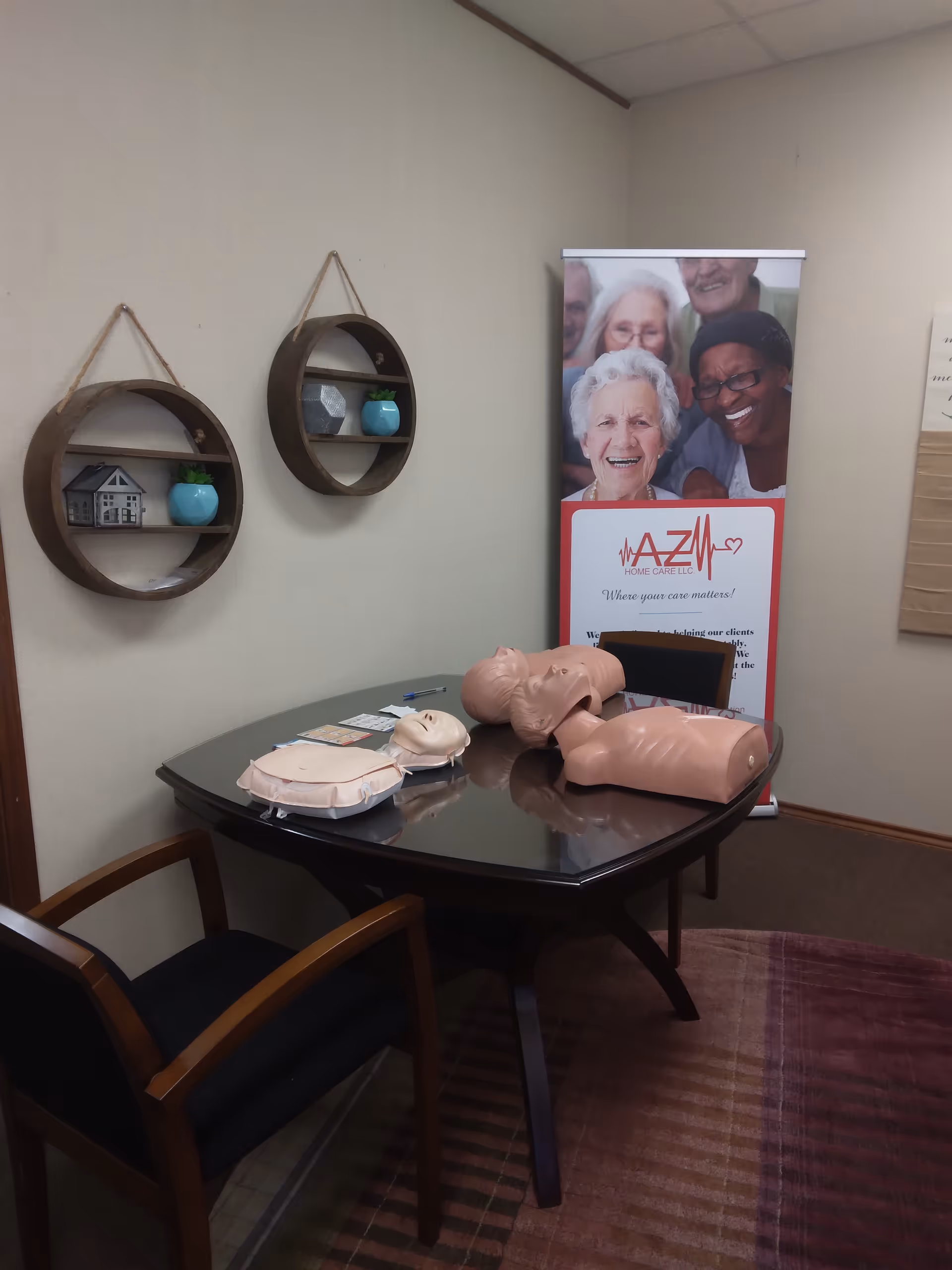 CPR mannequins on a table with an AZM Home Care banner inside a small conference room.
