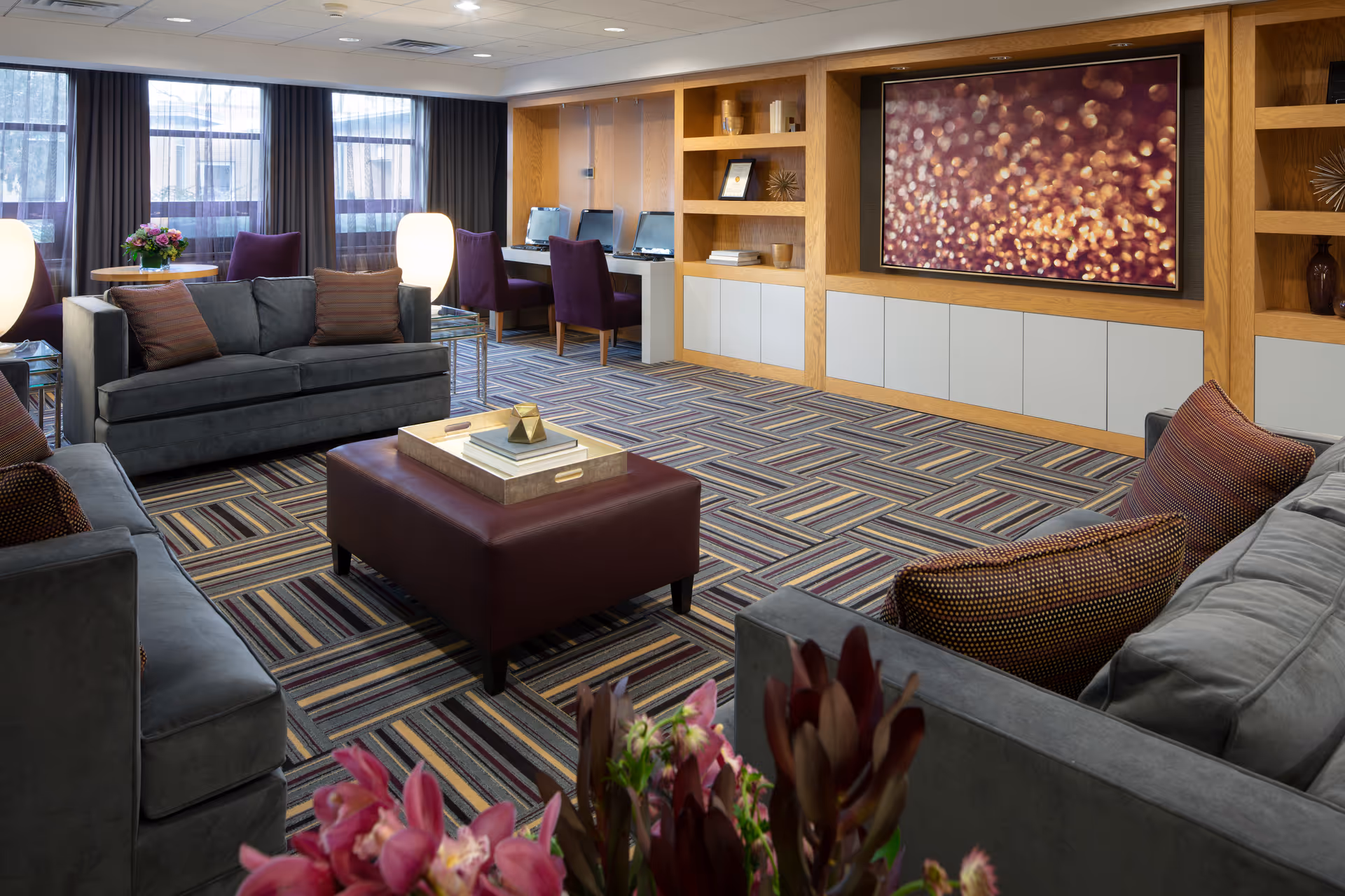 Spacious senior living lounge with gray sofas, patterned carpet, built-in shelves, and a large wall TV.