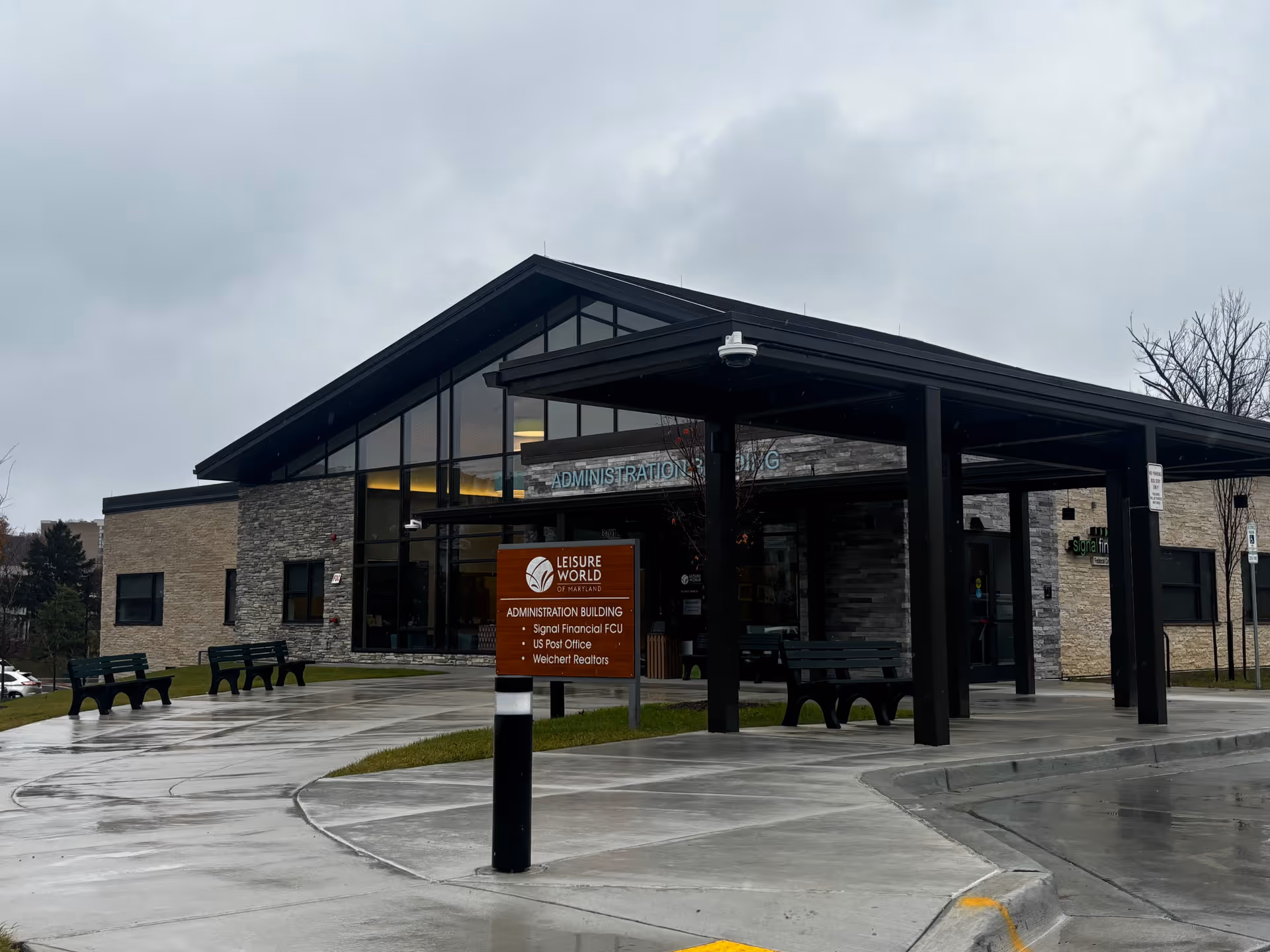 Front entrance of the Leisure World of Maryland administration building with a covered drop-off, sign, benches, and wet pavement under a cloudy sky.
