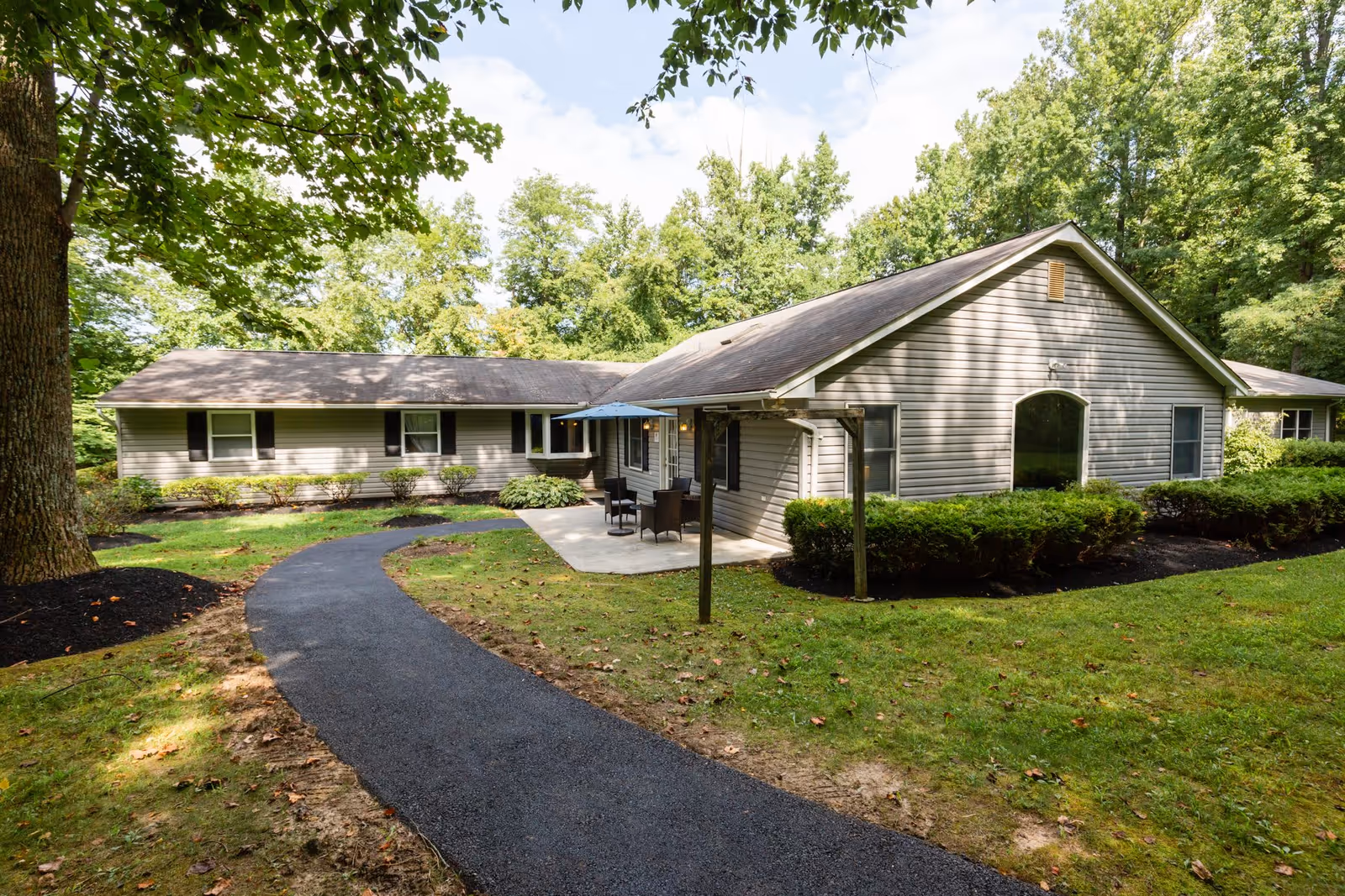 Single-story senior living building with a paved walkway leading to a patio with table and umbrella, surrounded by lawn and trees.
