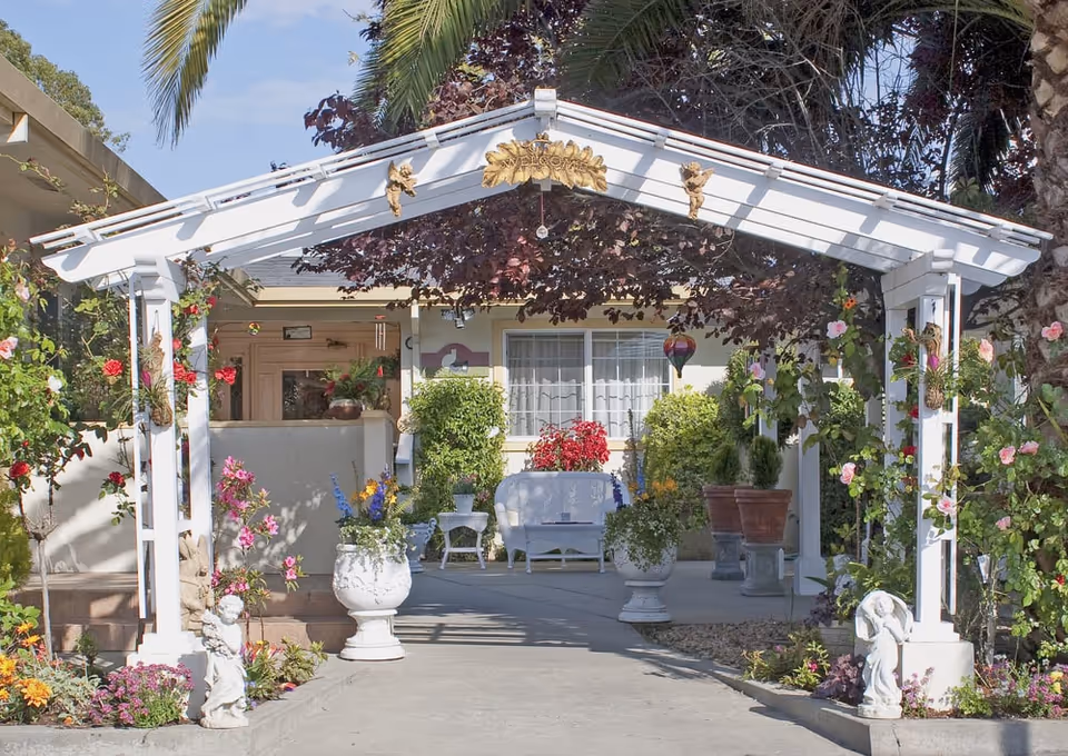 Front entrance of a small building framed by a white pergola, potted plants, flowers, and outdoor seating.
