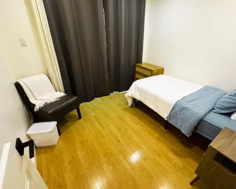 A compact bedroom with a single bed, chair, nightstand, and dark curtains.