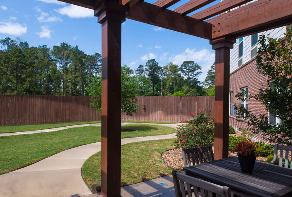 Patio with a pergola-covered outdoor dining table and chairs overlooking a curved sidewalk, lawn, and fenced yard beside a brick building.