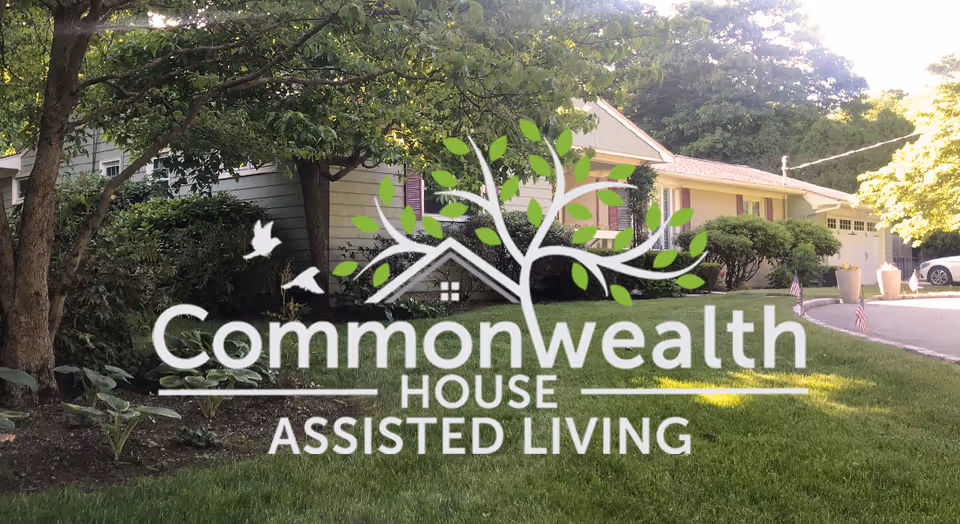 Exterior view of a residential-style assisted living facility with a well-maintained lawn, trees, shrubs, and a driveway. The building has light-colored siding with purple shutters and a garage. The logo for Commonwealth House Assisted Living is overlaid on the image.