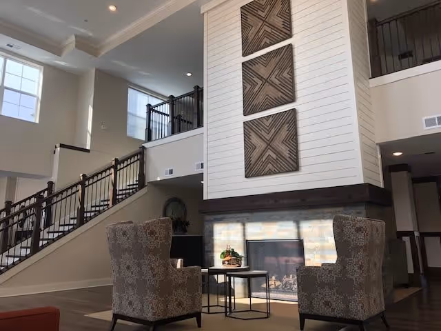 Bright two-story lounge with a central fireplace, two patterned armchairs, a small table, and a staircase leading to an upper balcony.
