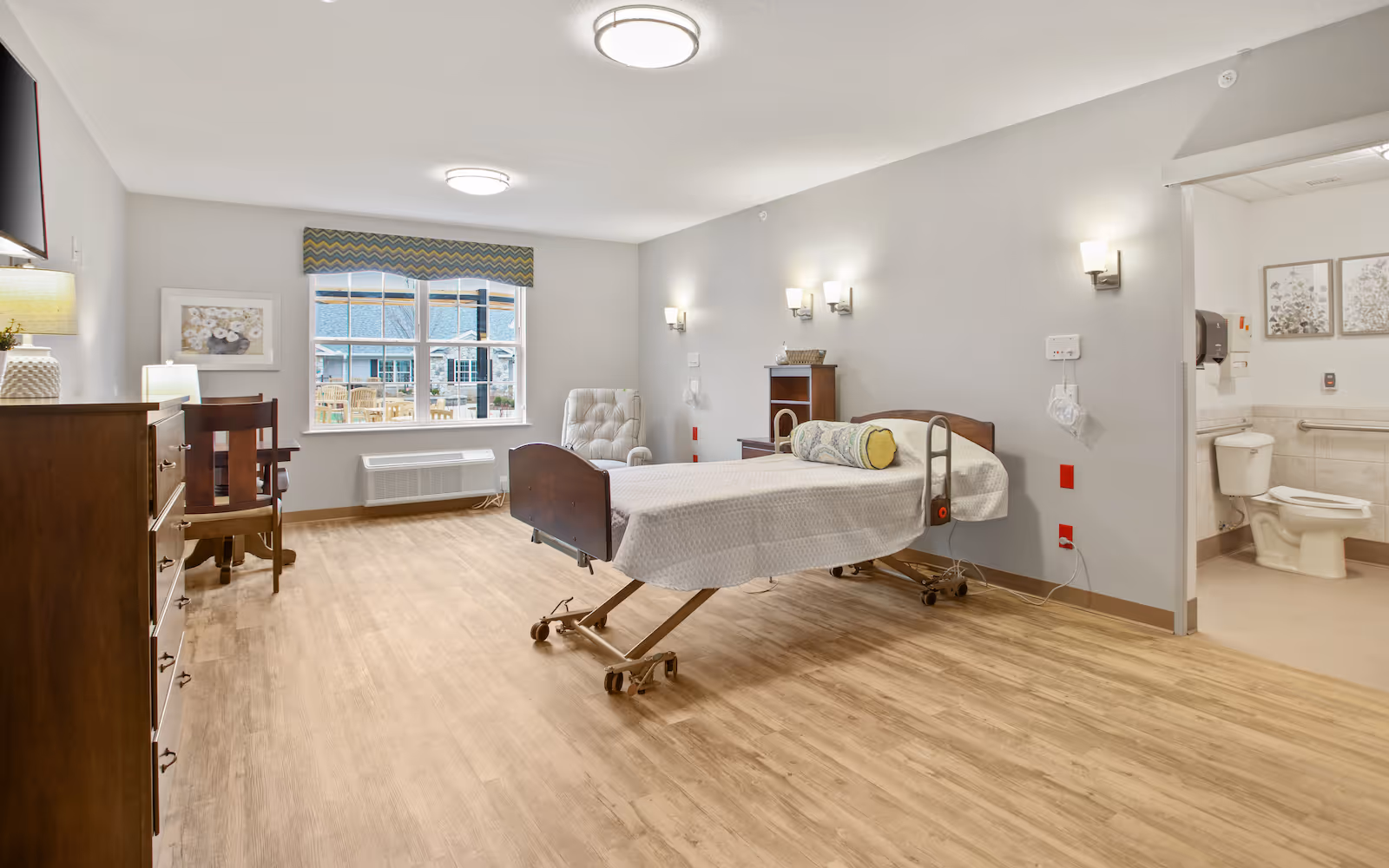 Well-lit senior living bedroom with a hospital-style bed, dresser, chair, window, and an adjoining bathroom.