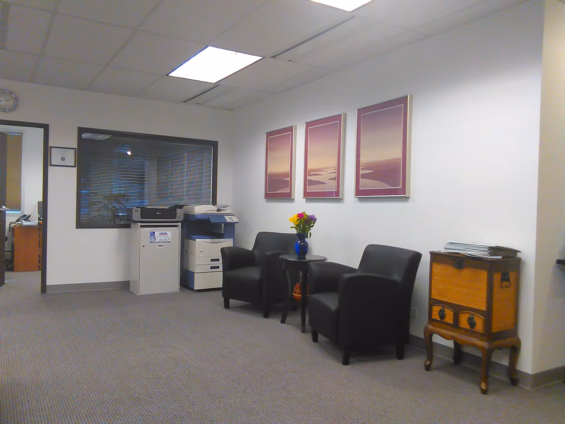 A waiting area with black chairs, a small table with flowers, wall art, and a printer.