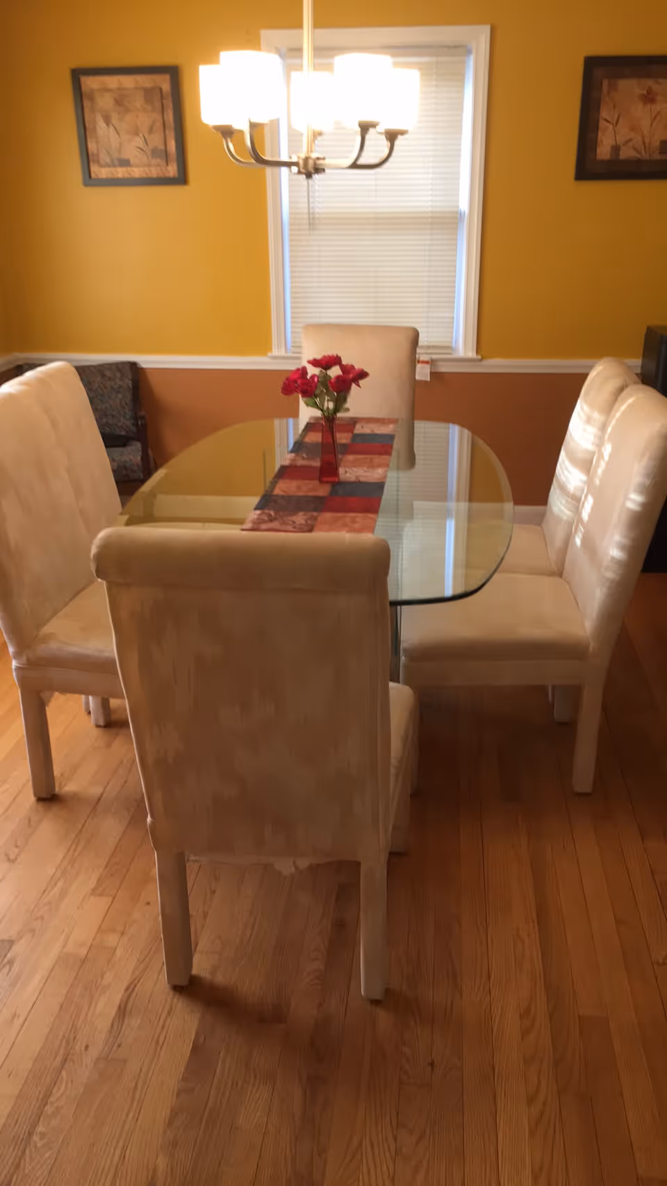 A dining room with a glass-top table surrounded by six beige upholstered chairs. A red vase with red flowers is placed on a multicolored table runner in the center of the table. The room has yellow and brown walls, a window with closed blinds, two framed floral paintings, and a chandelier hanging above the table.