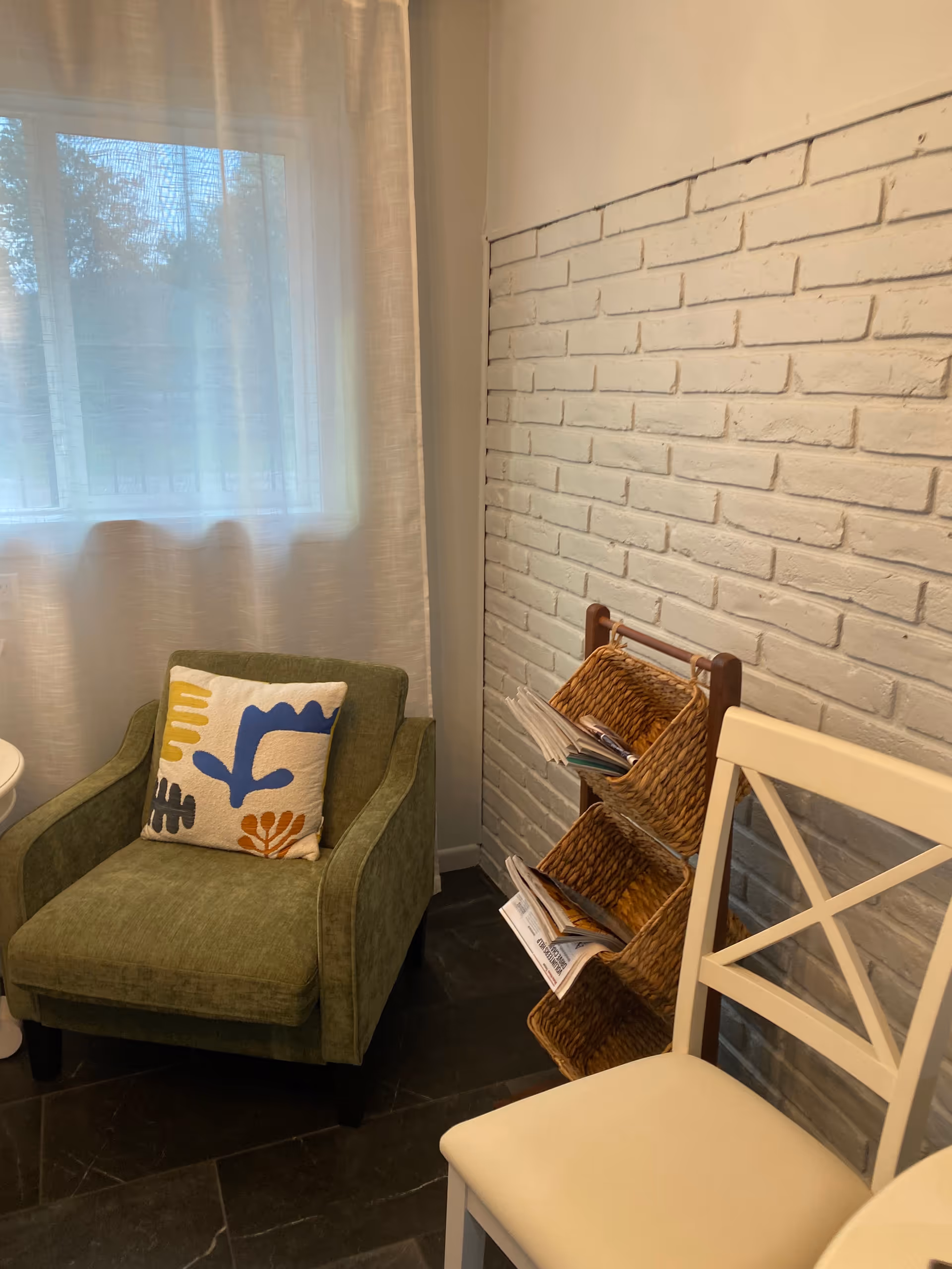 Green armchair with a decorative pillow beside a white chair and magazine rack near brick wall.
