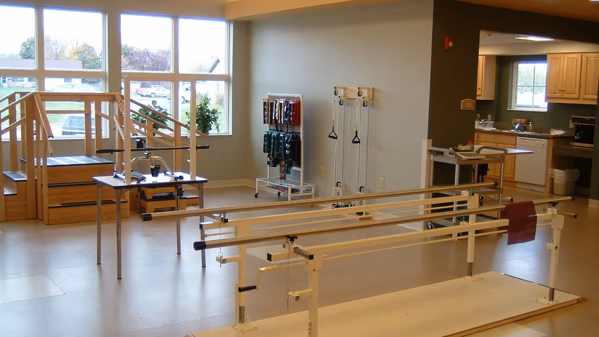 Interior therapy room with parallel bars, a practice staircase, mobility equipment and a kitchenette area in the background.