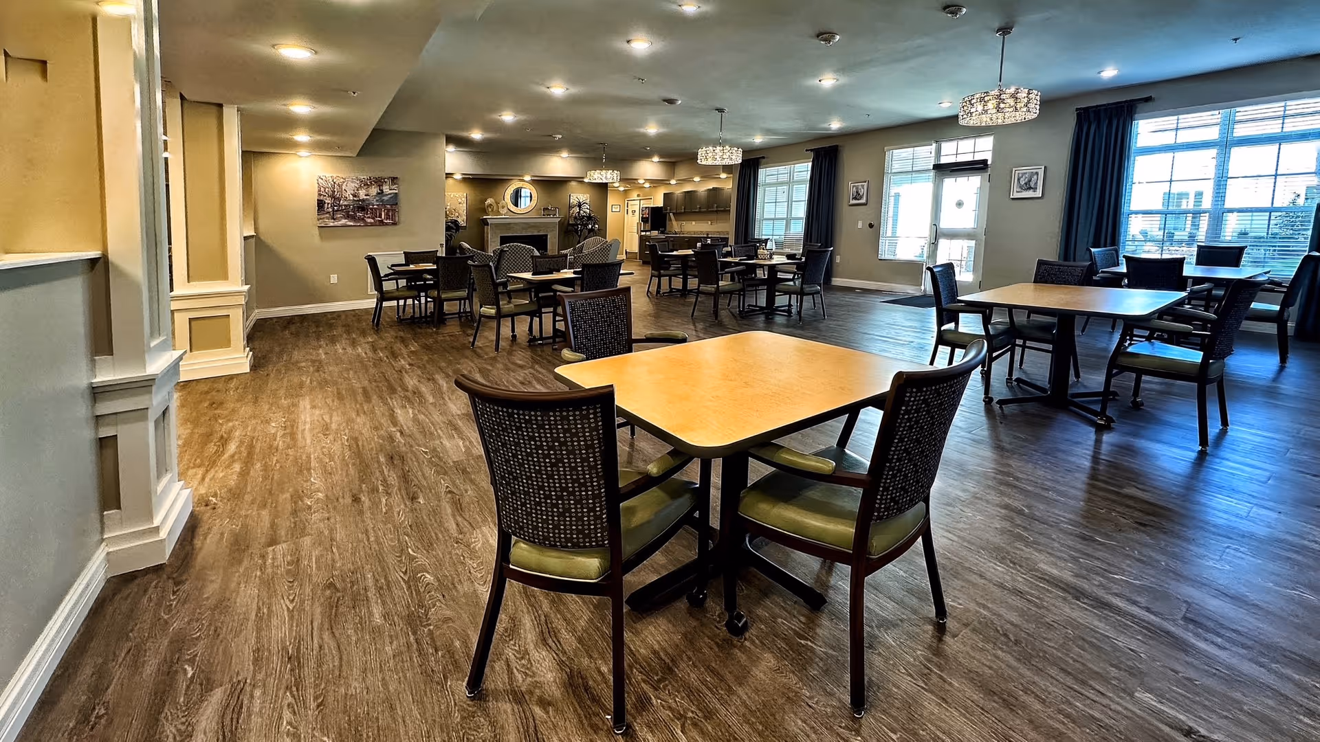 A spacious dining room with multiple wooden tables and chairs arranged neatly on a wood-patterned floor. The room features large windows allowing natural light to enter, a fireplace with a round mirror above it, and modern chandeliers hanging from the ceiling. The walls are painted in neutral tones with some framed artwork.