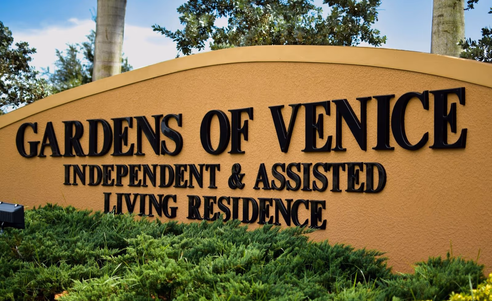 A large entrance sign reading 'GARDENS OF VENICE INDEPENDENT & ASSISTED LIVING RESIDENCE' with shrubs and palm trees behind it.