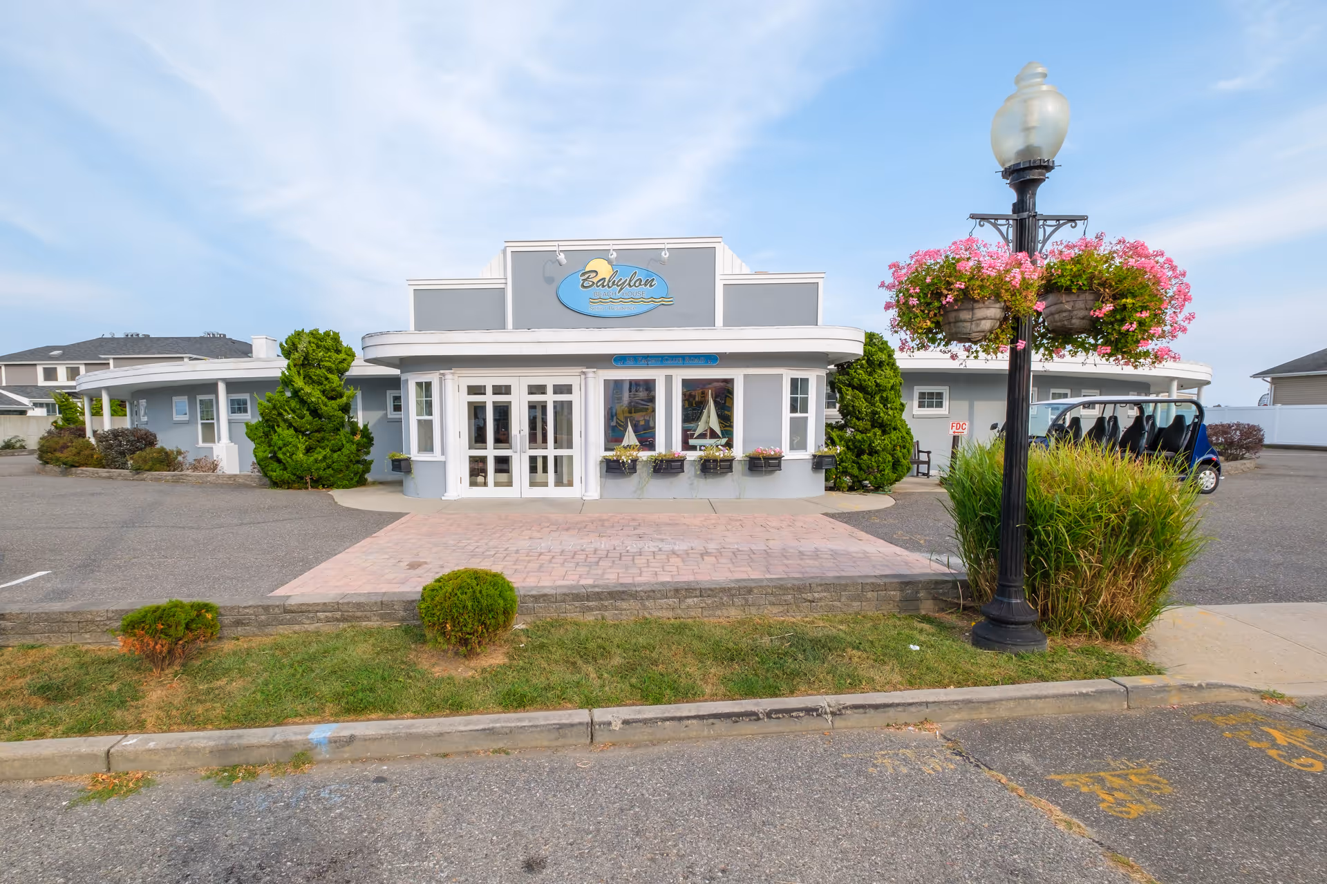 Front entrance of the Babylon Beach House building with a paved walkway, lamp post with hanging flower baskets, and surrounding parking area.