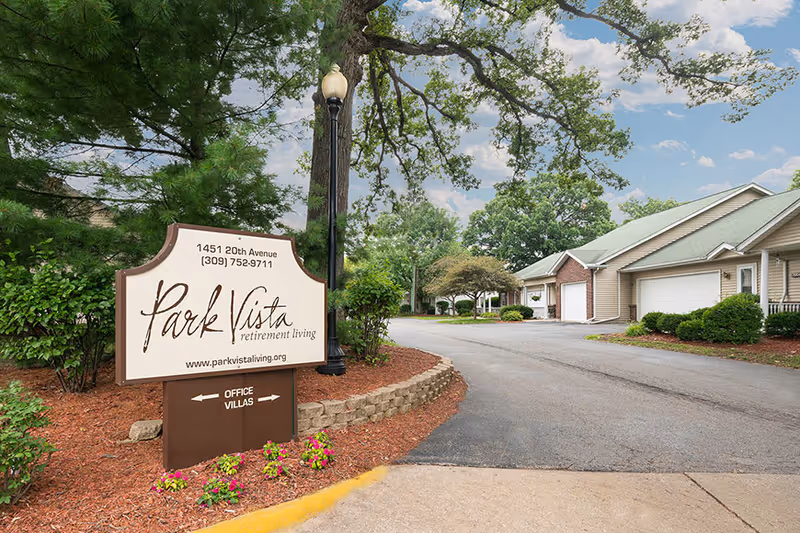 Entrance sign for Park Vista retirement living located at 1451 20th Avenue with phone number and website, surrounded by landscaping with flowers and bushes, a paved driveway leading to residential buildings with garages, and trees under a partly cloudy sky.